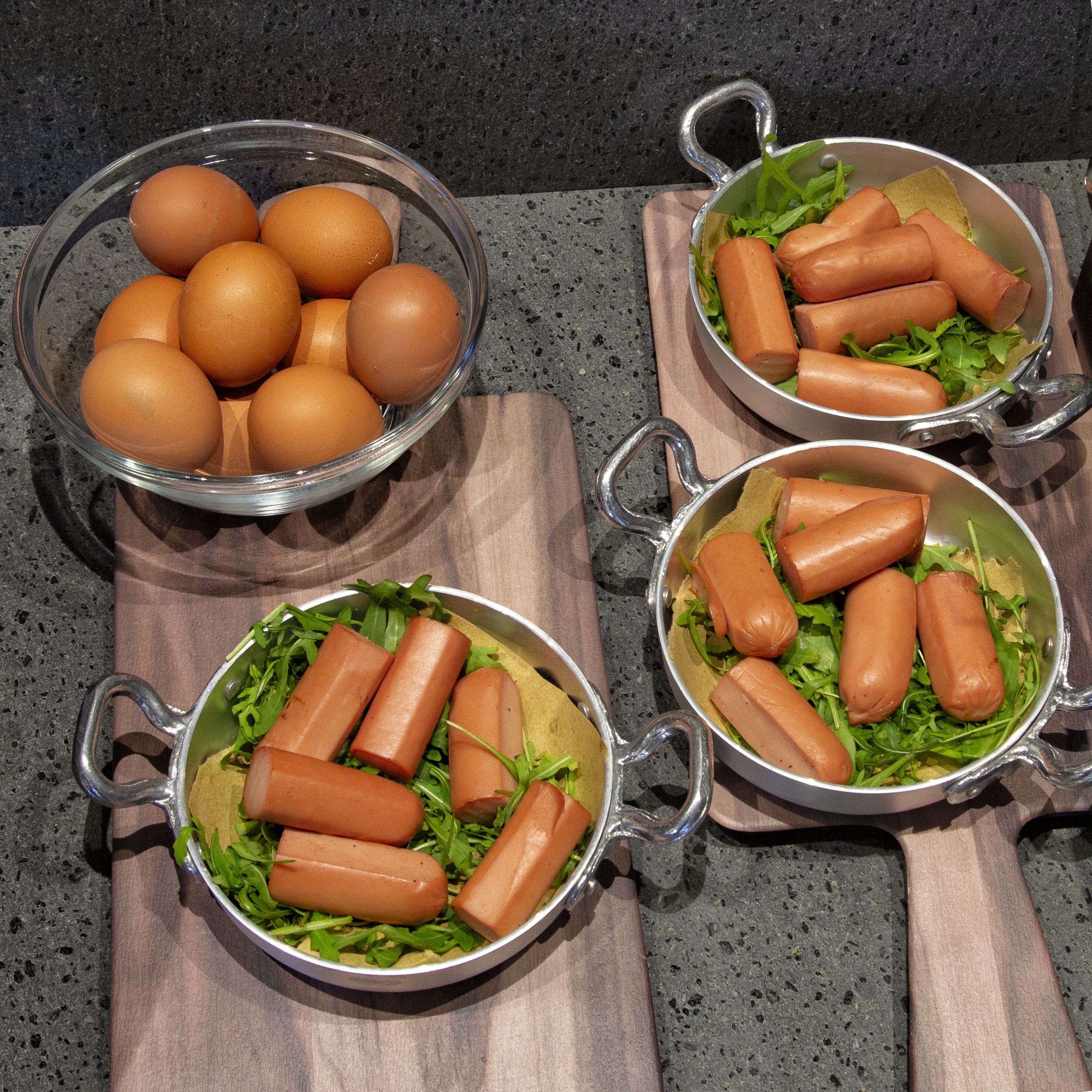 Uma bancada de cozinha com três tigelas contendo ovos e cachorros-quentes sobre uma tábua de corte.