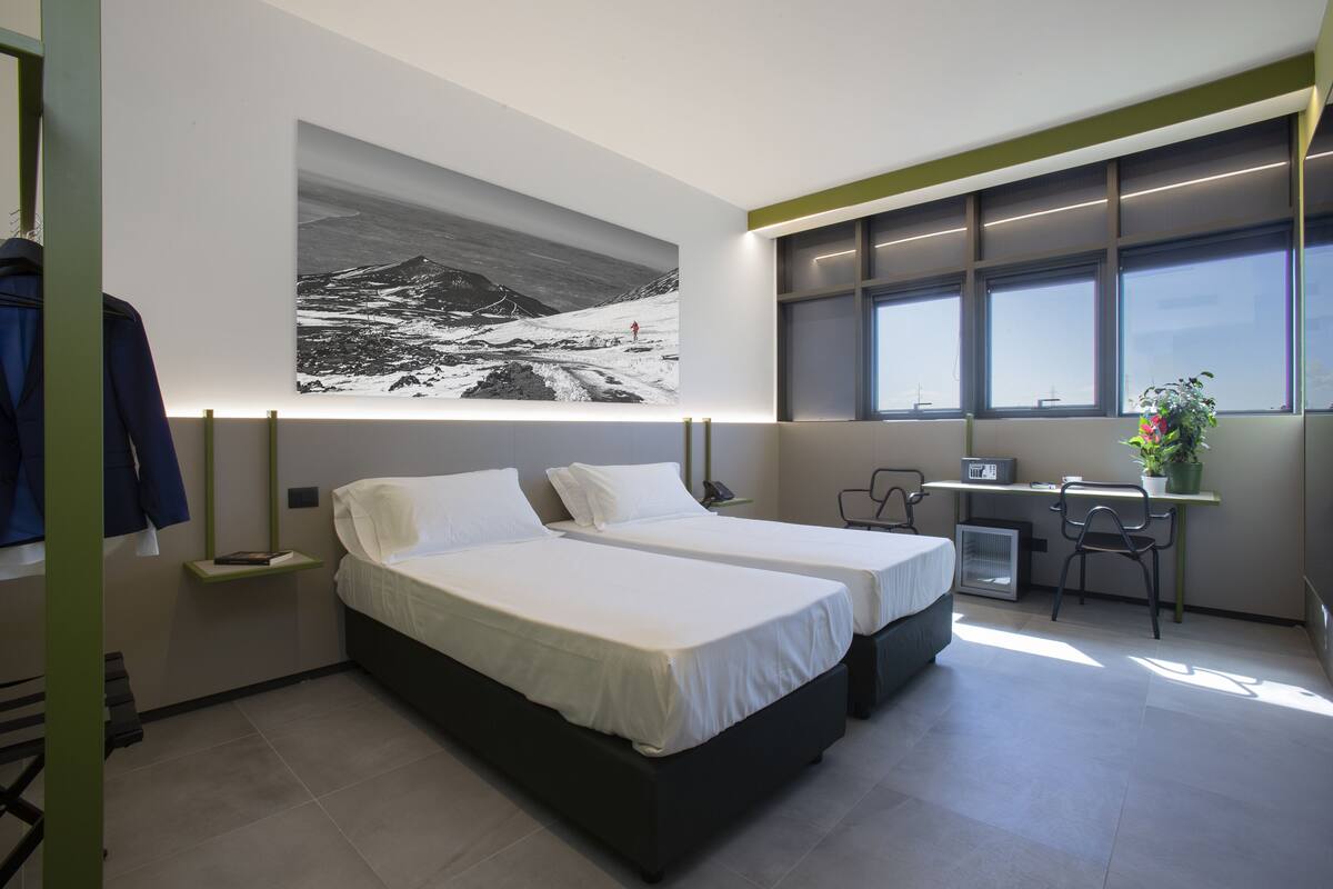 A modern hotel room with two beds, a desk, a chair, a large window, and a wall-mounted artwork.