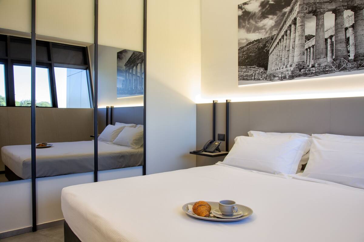 A hotel room with two beds, a breakfast tray with croissant and coffee, a phone on a nightstand, and a large framed picture of ancient columns.