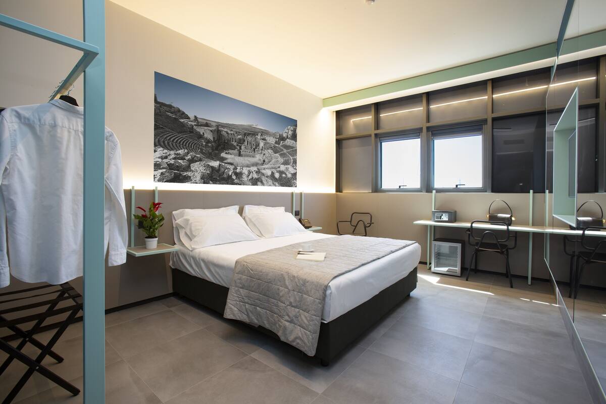 A modern hotel room with a large bed, a desk, and a view of a mountainous landscape.