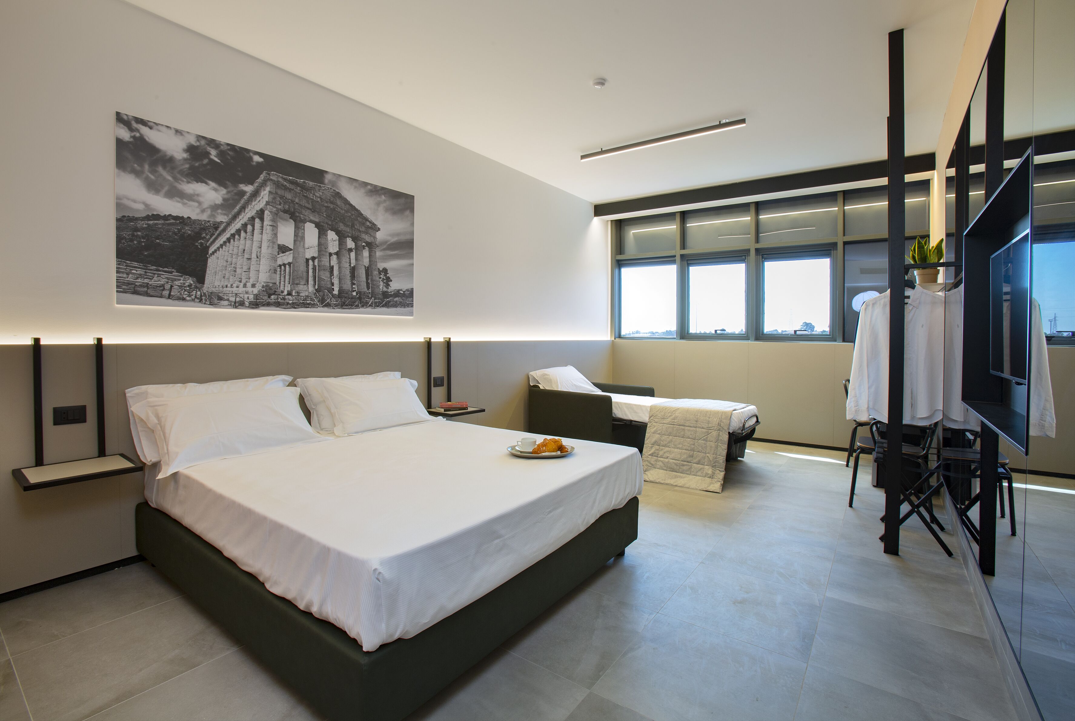 Una habitación de hotel moderna con una cama grande, un sofá pequeño, un televisor y una ventana con vista a la ciudad.