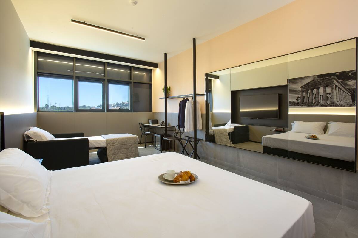 A modern hotel room with a large bed, a desk, a chair, a TV, and a large window offering a city view.