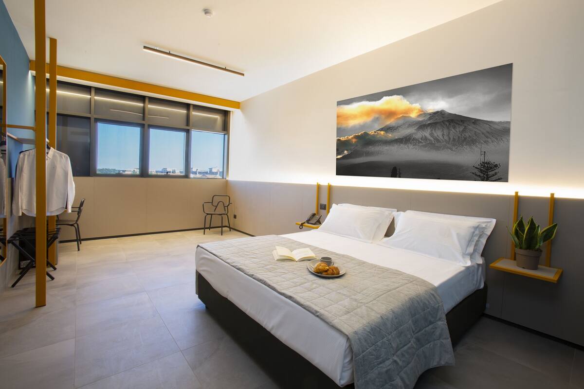 A modern hotel room with a large bed, a bedside table with a lamp, a window with a city view, and a painting on the wall.