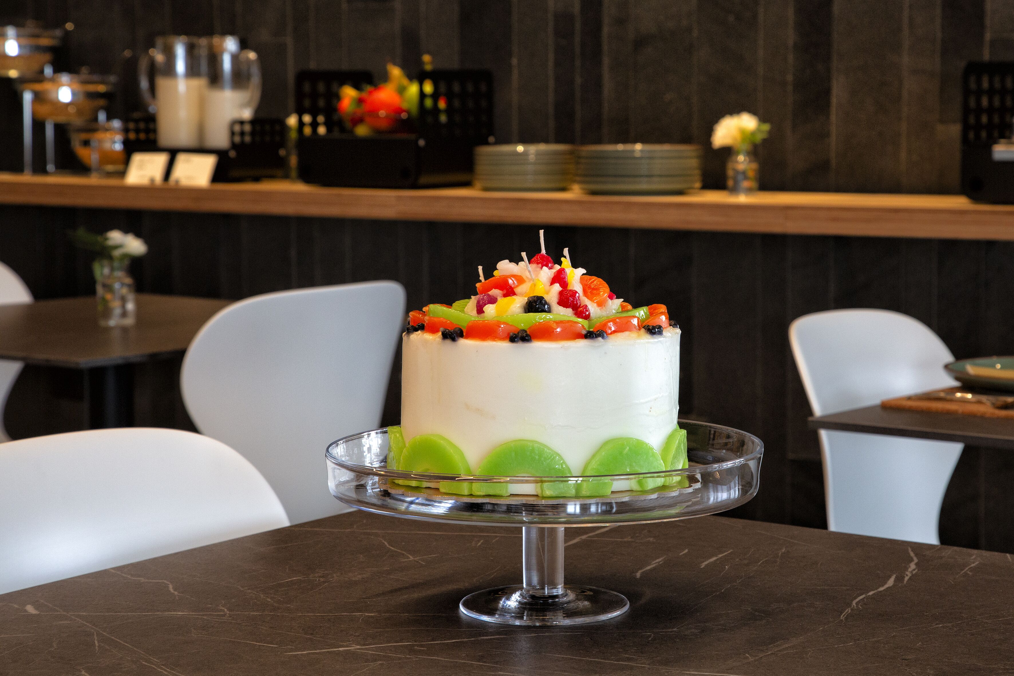 A cake on a glass stand in a dining area with chairs and a counter in the background.