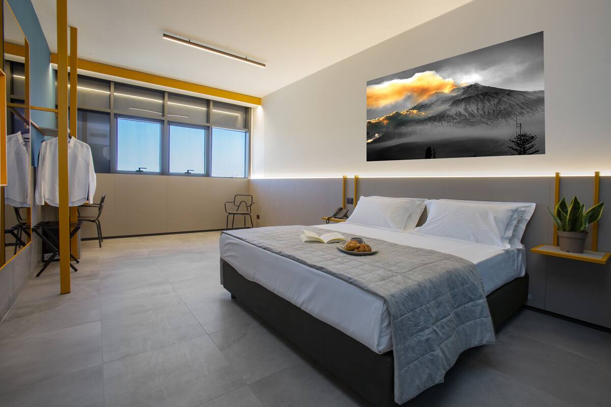 A modern bedroom with a large bed, a window with a view, a painting on the wall, and a small table with a plant.
