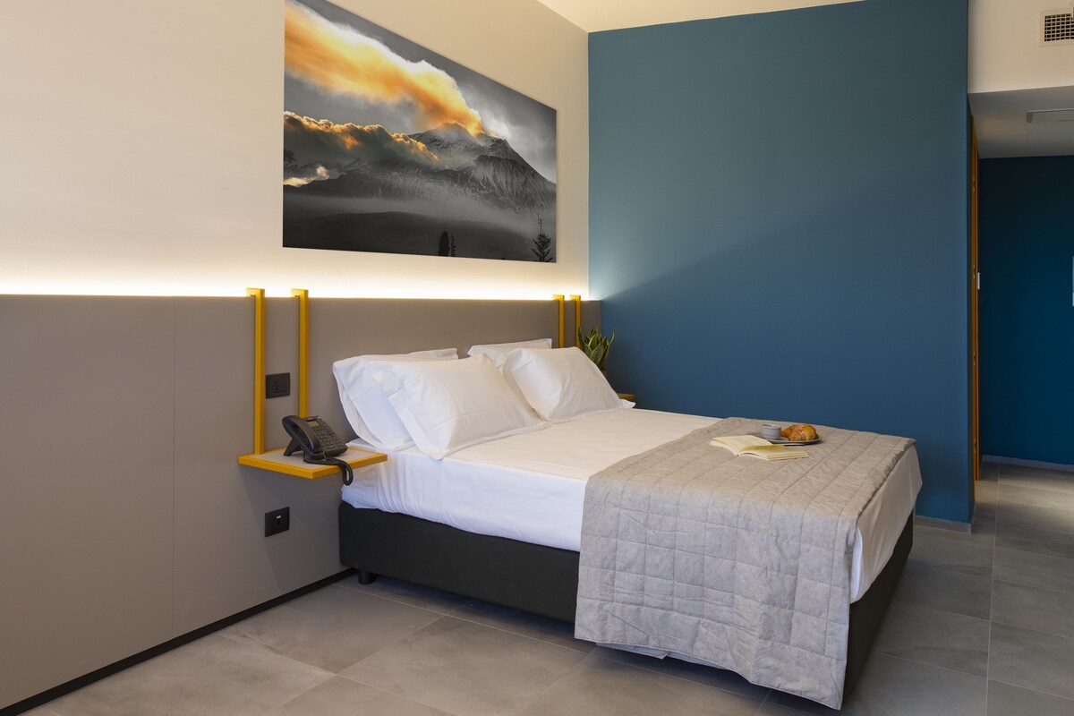 A modern hotel room with a large bed, bedside table with a phone, and a painting of a mountain landscape.