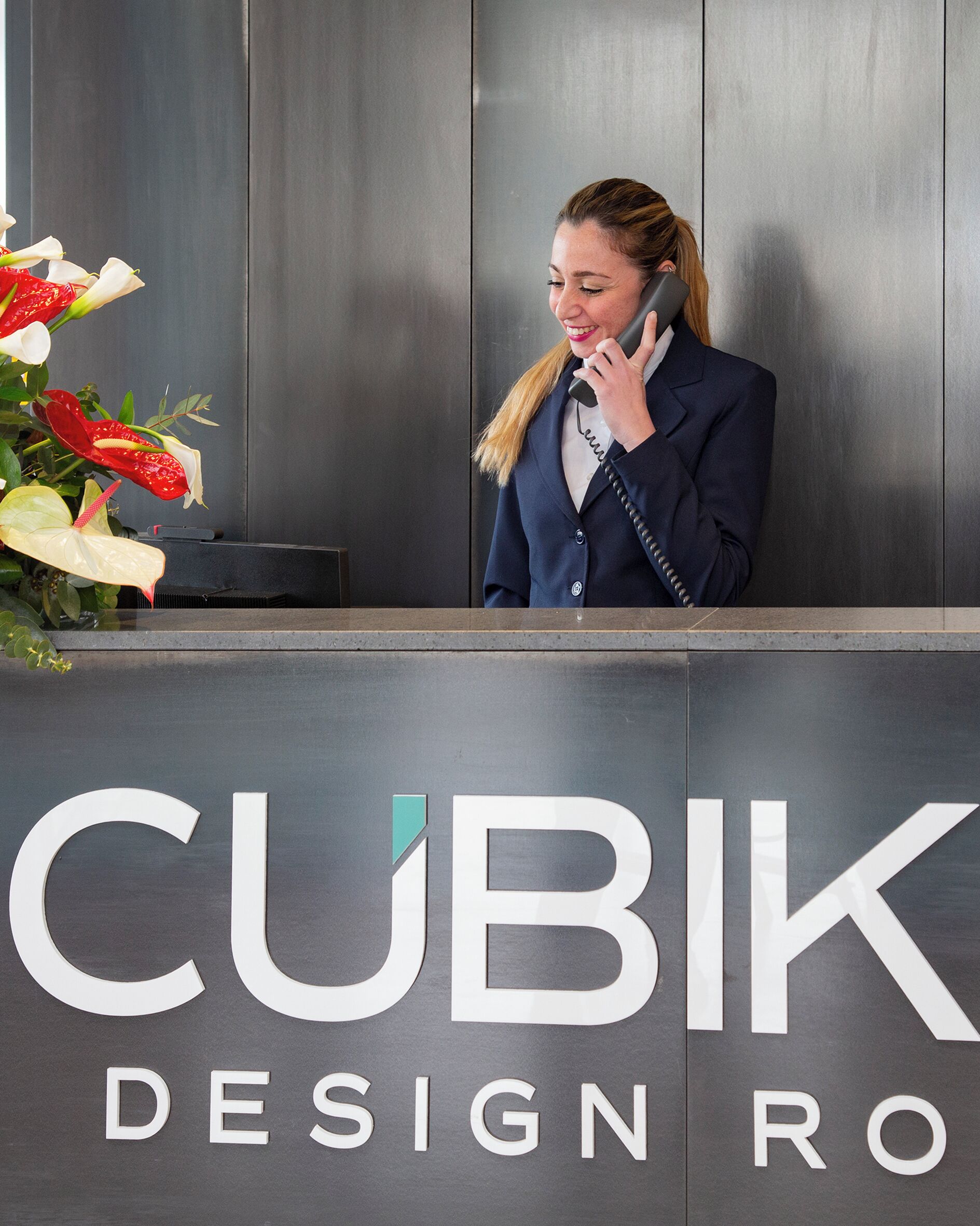 Ein Empfangsschalter mit einem Schild, auf dem 'CUBIK DESIGN RO' steht, und eine Frau am Telefon.