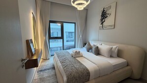 A hotel room with a large bed, a view of buildings outside, a modern light fixture, and a painting on the wall. - Liva Suites By Hostiva - Dubai South (Dubai)