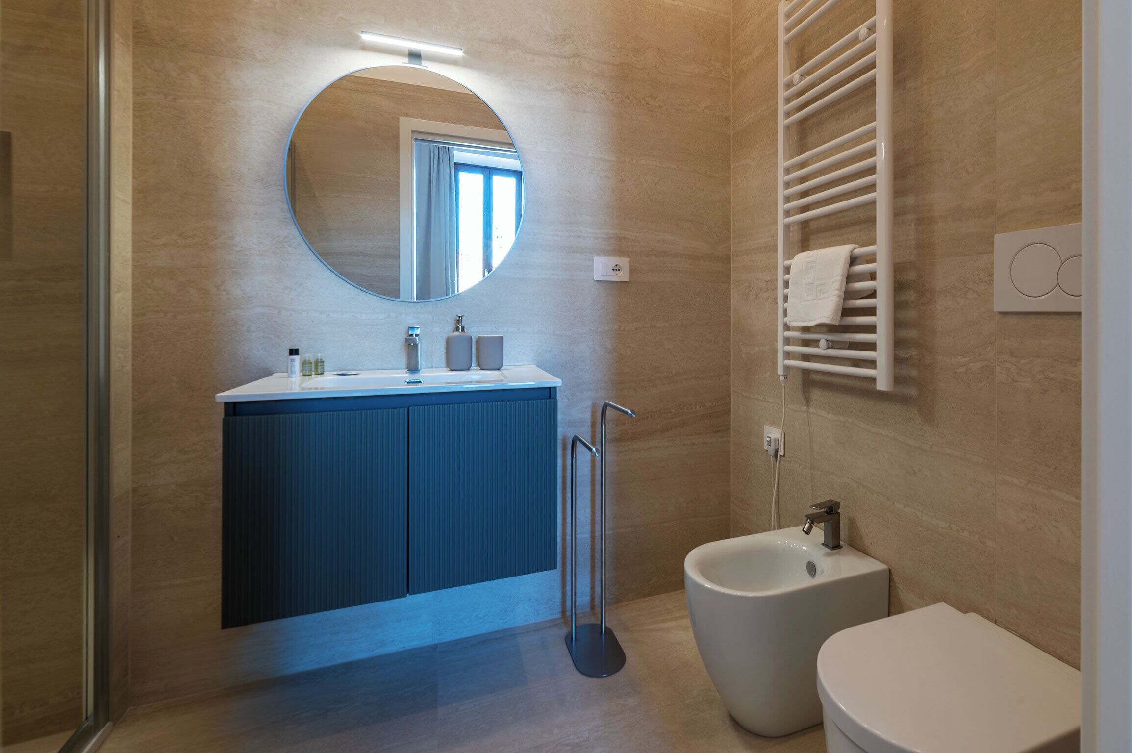 A modern bathroom with a round mirror, a sink with a blue cabinet, a bidet, and a towel rack.