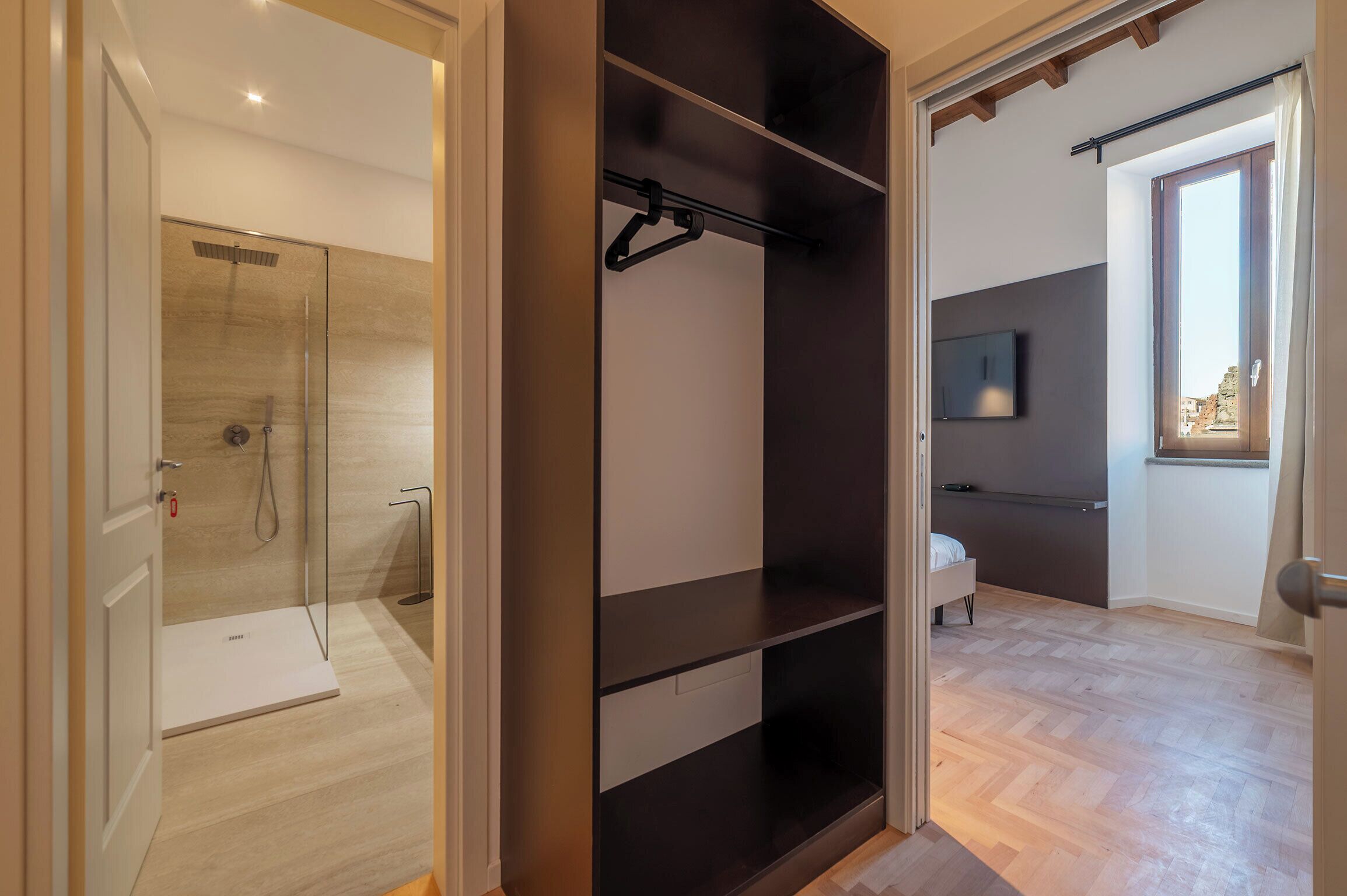 Un baño moderno con ducha de vidrio, un armario negro con barra para colgar ropa y una cama en la habitación contigua.