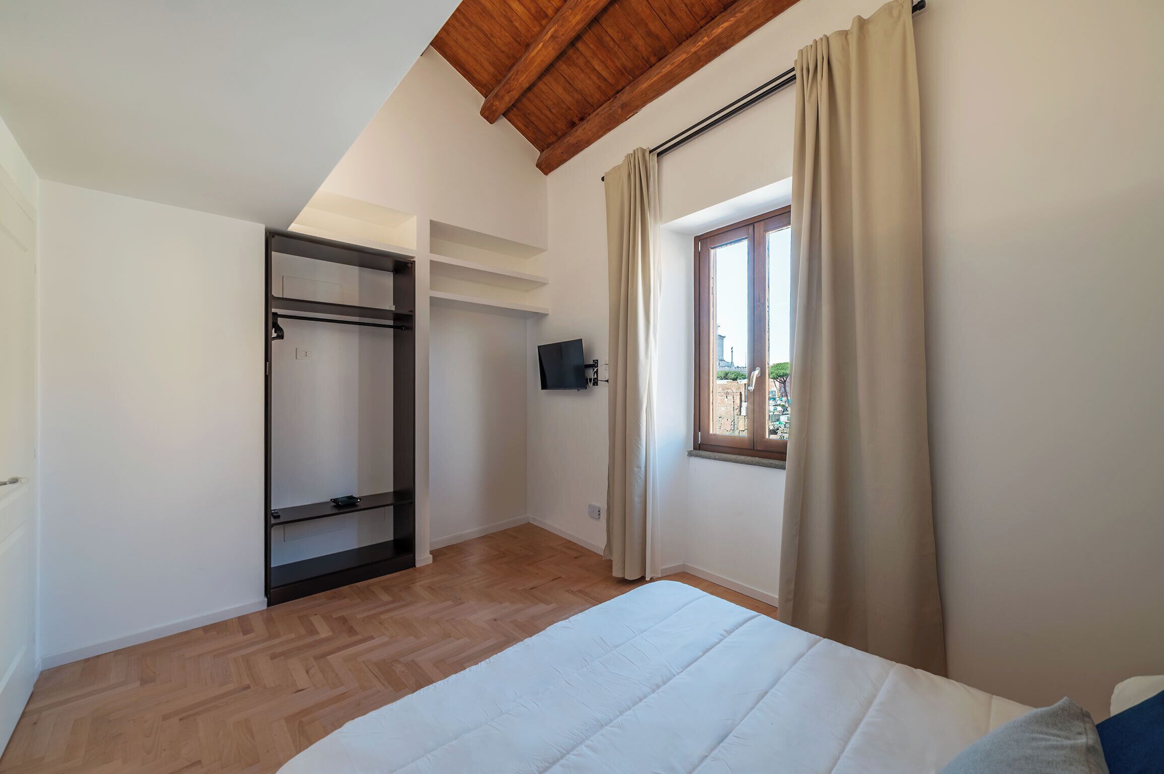 Un dormitorio con cama, suelo de madera, ventana con cortinas, televisor y armario.