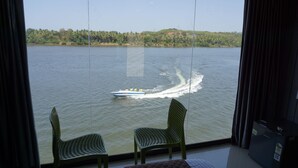 Vista desde una ventana sobre un lago con un bote dejando una estela, dos sillas verdes y un vistazo a un edificio al otro lado del agua.