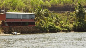 Edificio de dos pisos con fachada roja y un balcón, ubicado junto a una masa de agua con palmeras al fondo.