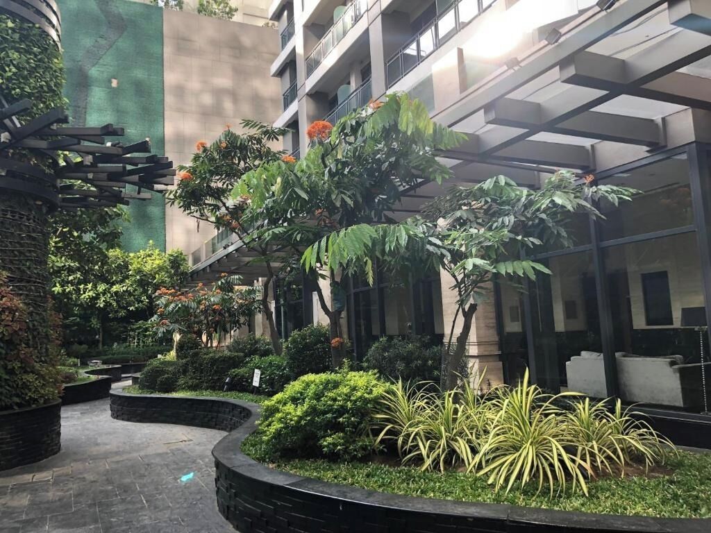 A courtyard with a curved pathway, surrounded by various plants and trees, and a building with balconies in the background.