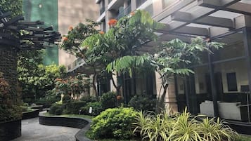 A courtyard with a curved pathway, surrounded by various plants and trees, and a building with balconies in the background.