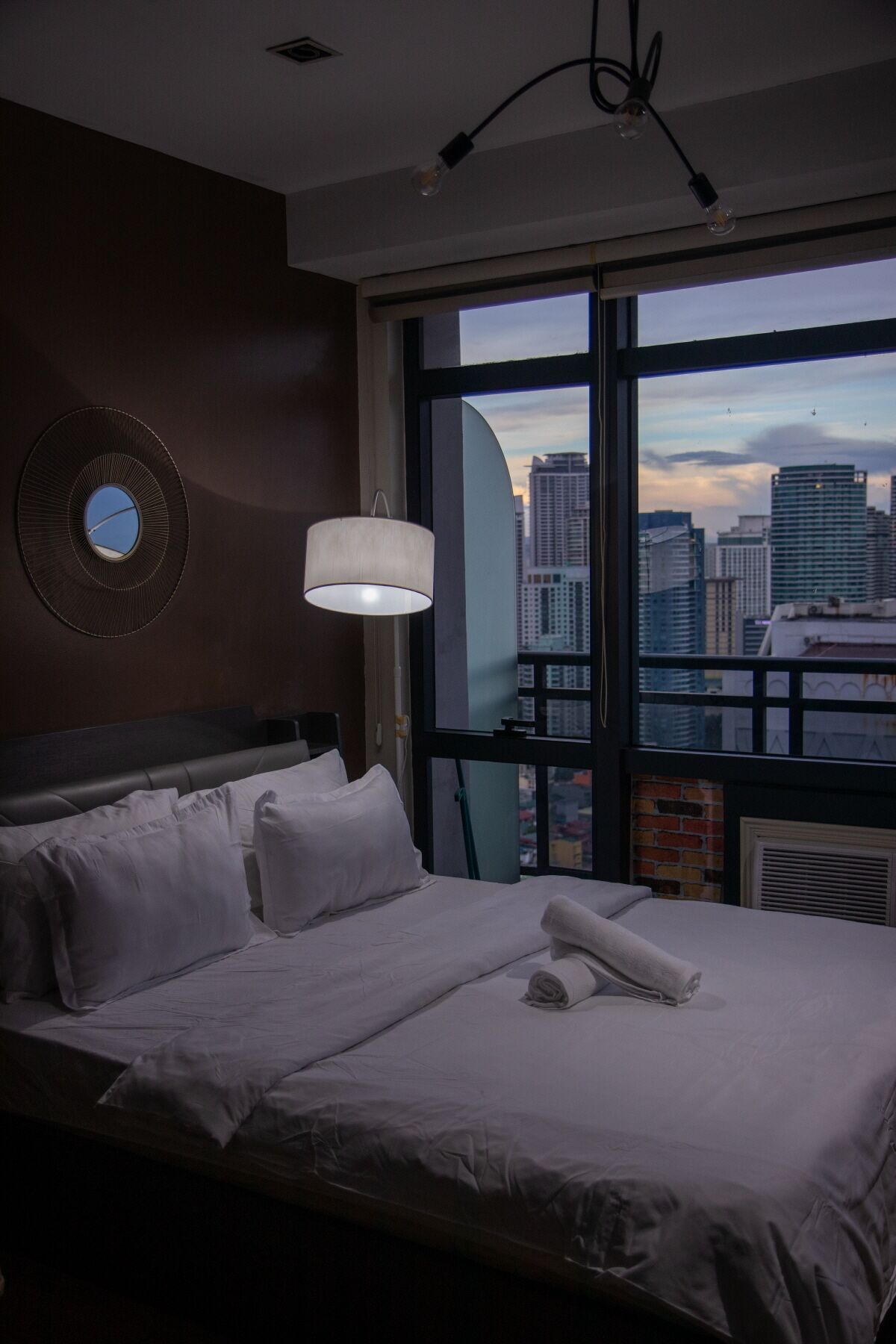 A hotel room with a large bed, a round mirror on the wall, and a view of a cityscape through a large window.