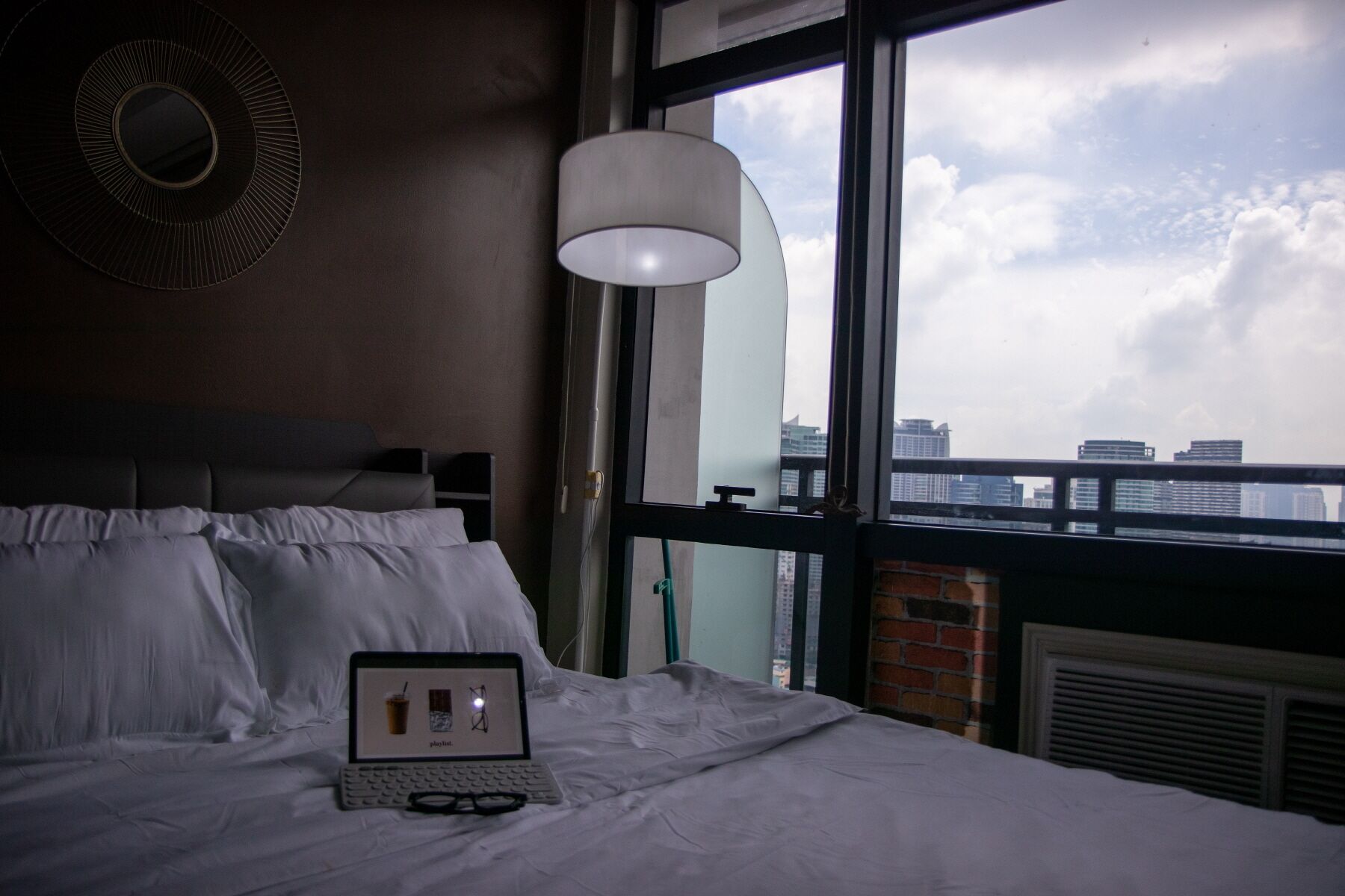 A hotel room with a bed, a laptop, and a view of the city through large windows.