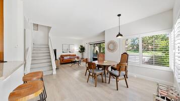 A spacious living area with a staircase, a dining table with chairs, a sofa, and a large window with blinds.