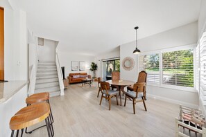 A spacious living area with a staircase, a dining table with chairs, a sofa, and a large window with blinds.