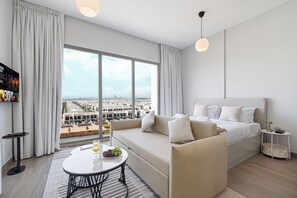 Una camera d'albergo moderna con un letto grande, un divano, un tavolo da pranzo con della frutta e vista sulla città attraverso ampie finestre.