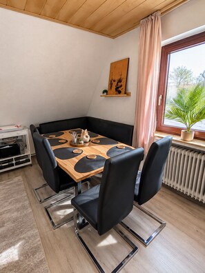 A dining area with a wooden table, black chairs, a black cushioned bench, a painting on the wall, a window with a plant, and a radiator. - "Meer" Vacation Rental - with a terrace and garden (Butjadingen)