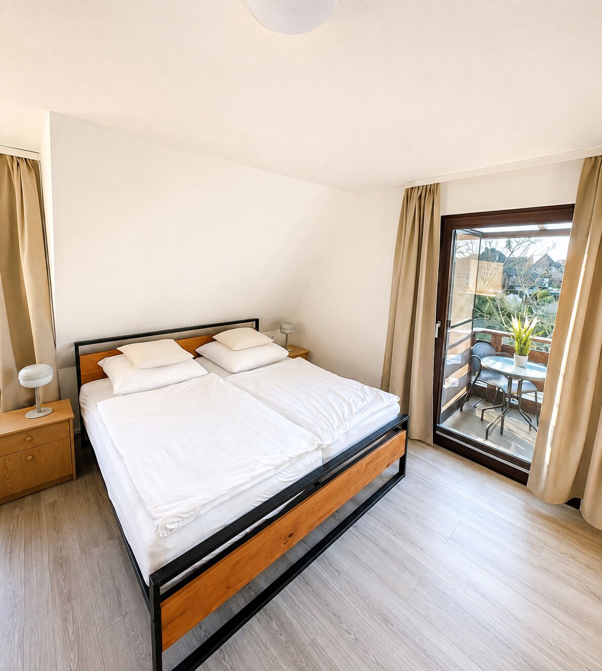 A double bed with a wooden frame and white bedding, a bedside table with a lamp, and a view of a balcony with outdoor furniture.
