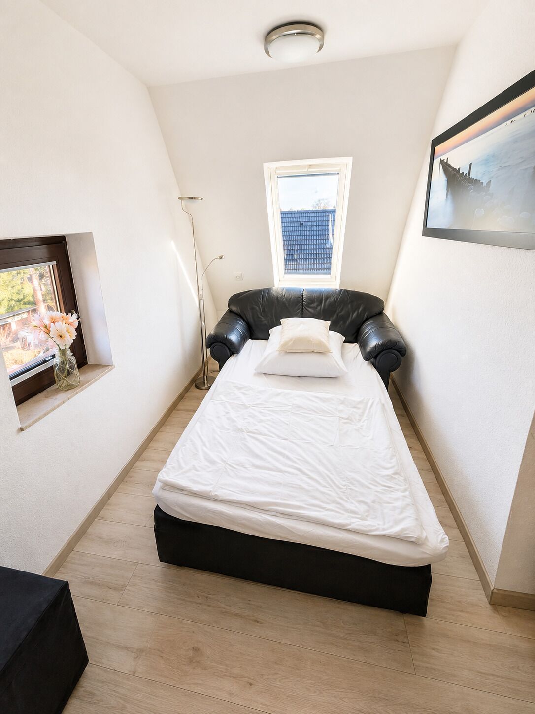 Une petite chambre avec un lit simple, un plafond en pente, une fenêtre, un lampadaire et un tableau encadré au mur.