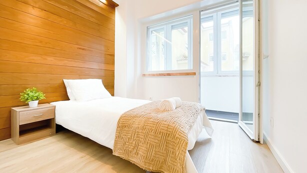 Ein Schlafzimmer mit Holz-Kopfstück, einem Bett mit weißer Bettwäsche, einem Nachttisch mit Pflanze und Blick durch ein großes Fenster.