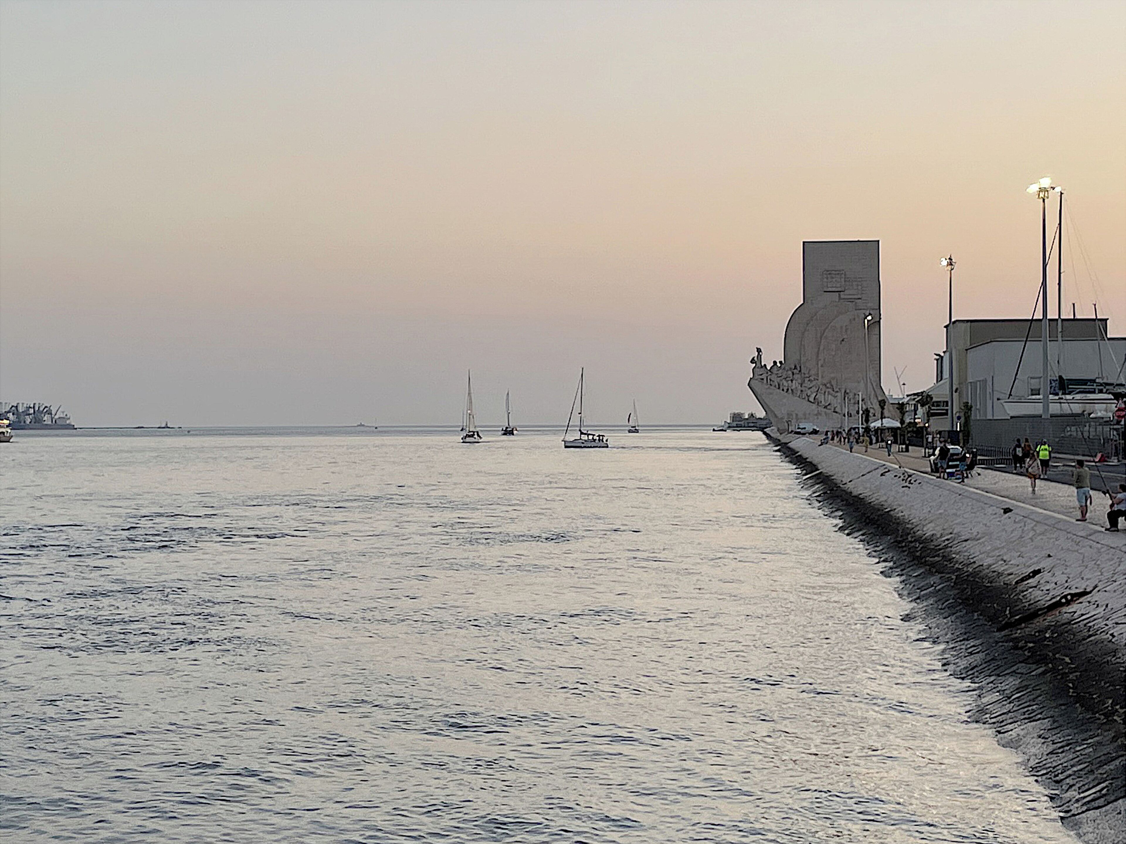 A waterfront promenade with a large building, a promenade with people, and sailboats on the water.