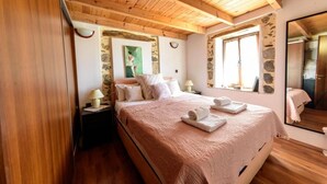 Ein Schlafzimmer mit Holzbalkendecke, Steinwänden, einem Bett mit rosa Bettdecke, zwei Nachttischlampen und einem großen Spiegel.