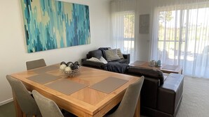 A modern living room with a wooden dining table, grey upholstered chairs, a dark brown sofa, and a large abstract painting on the wall. - Bay & wetland views in beautiful Williamstown, Relax, unwind and enjoy (Williamstown)
