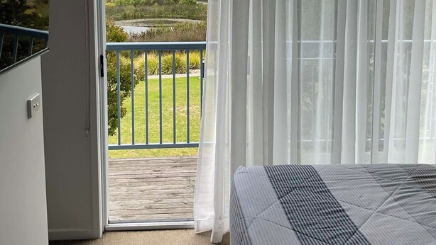 A bedroom with a bed, a balcony with a view of a grassy area and trees, and sheer curtains. - Bay & wetland views in beautiful Williamstown, Relax, unwind and enjoy (Williamstown)