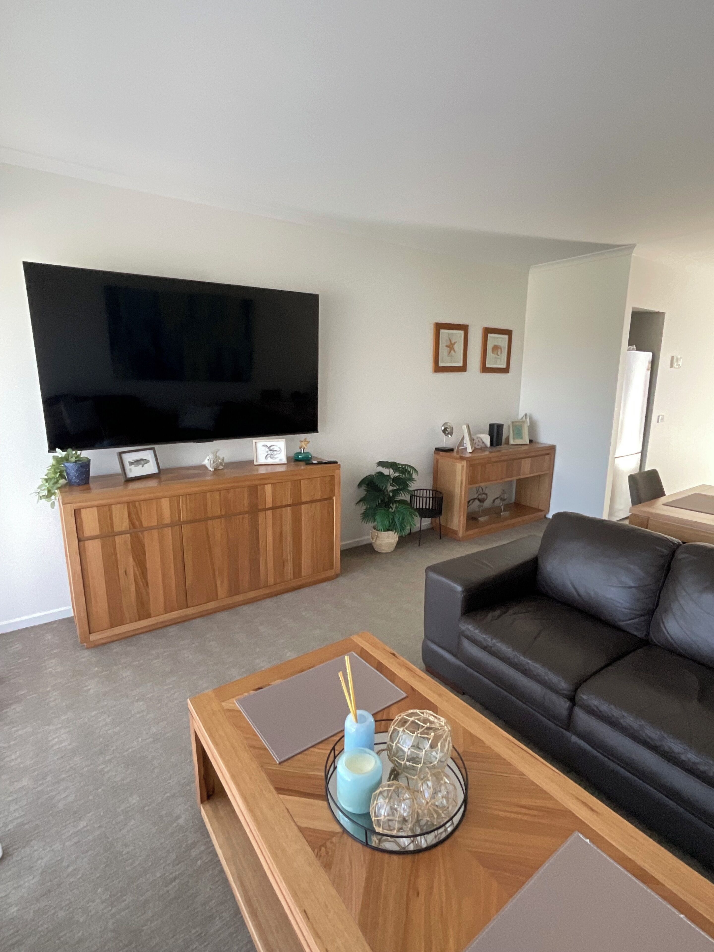 A living room with a large flat-screen TV, wooden entertainment unit, black leather sofa, and a coffee table with decorative items.