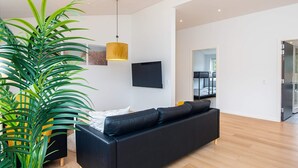 Uma sala de estar moderna com um sofá preto, uma planta verde grande, uma luminária pendente amarela e uma TV de tela plana fixada na parede.