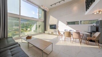 A modern living room with a large window, a flat-screen TV, a dining area with chairs, and a kitchen area with a refrigerator.