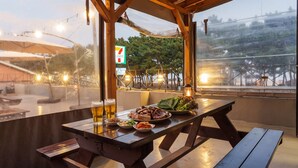 Un espace de restauration en plein air avec vue sur un pont et des arbres, une enseigne 7-Eleven, et une table en bois avec de la nourriture et des boissons.