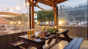 An outdoor dining area with a view of a bridge and trees, a 7-Eleven sign, and a wooden table with food and drinks. - Taean Sagye Pension (Taean)