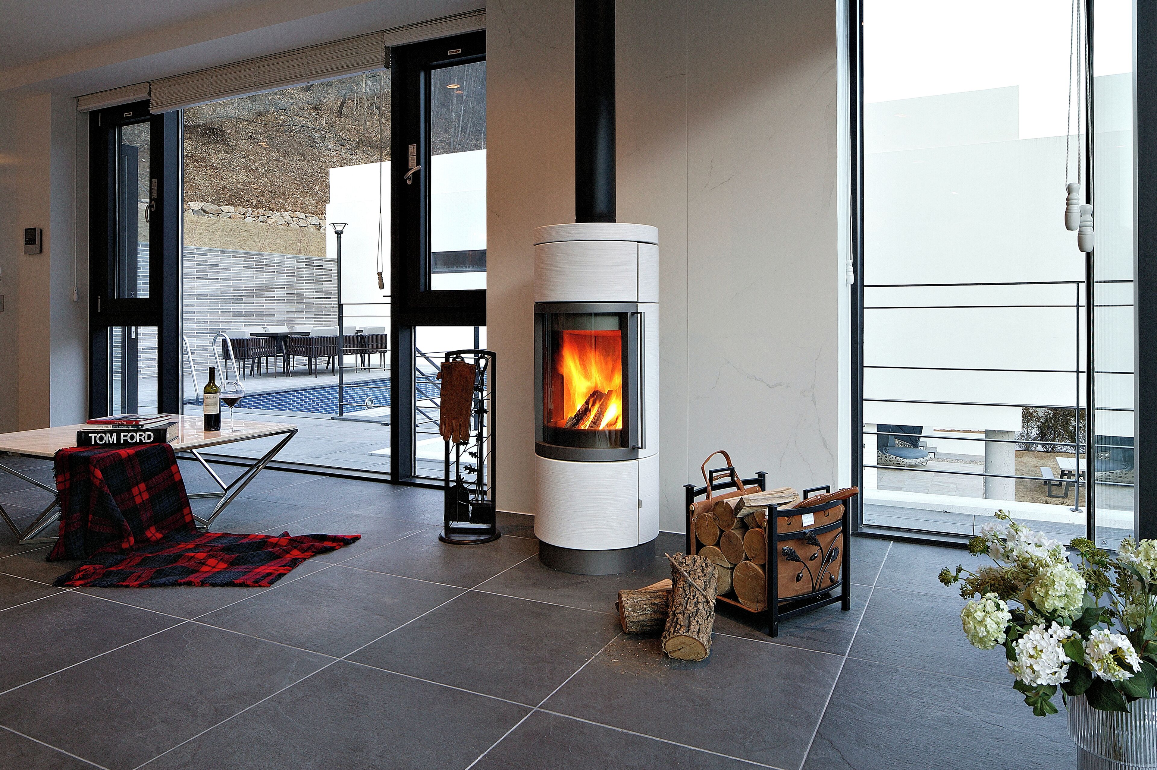 A modern living room with a wood-burning stove, a dining area with a table and chairs, and a large window offering a view of the outdoors.