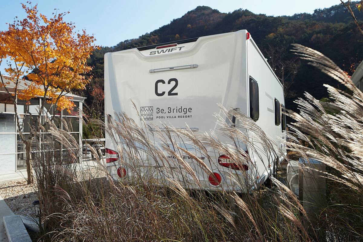 A white RV with the label "C2" and "Be, Bridge POOL VILLA RESORT" parked near a building with a glass facade.