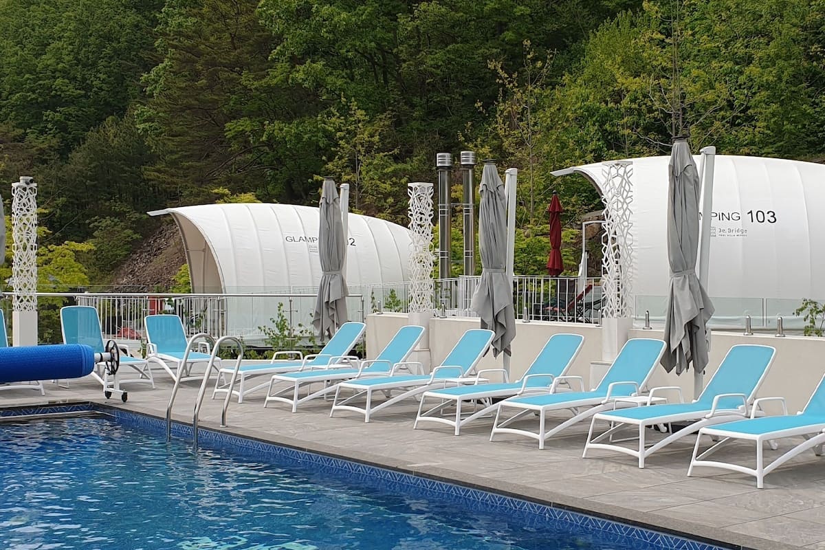 A swimming pool with blue lounge chairs and umbrellas, surrounded by lush greenery and a hill in the background.