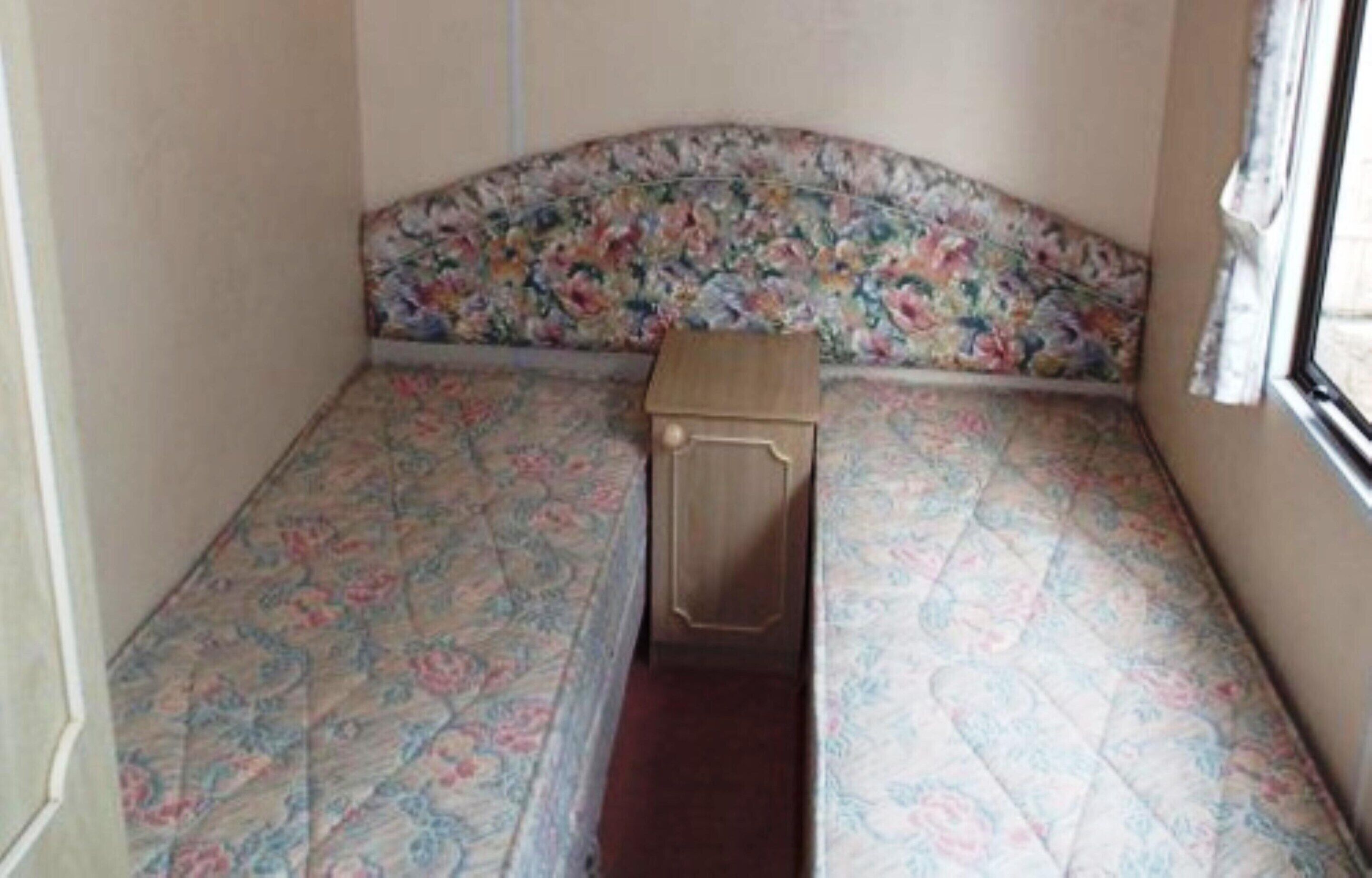 A small room with a floral-patterned bedspread, a wooden nightstand, and a window with curtains.