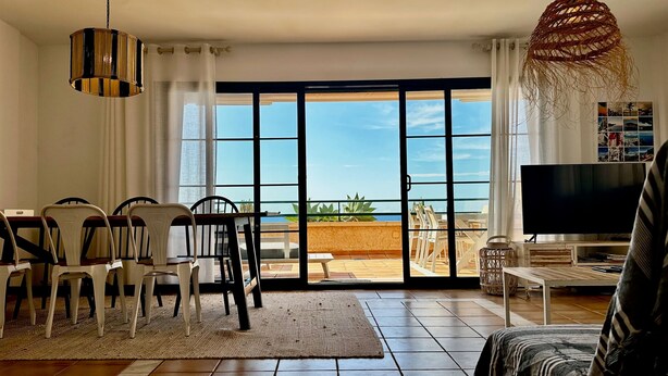 Een ruime woonkamer met een grote schuifpui naar een balkon met uitzicht op zee. Er is een eethoek met stoelen en een tafel, een televisie en een geweven lamp.