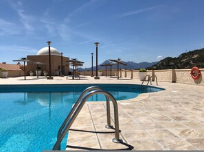 A swimming pool with a metal ladder, surrounded by lounge chairs and umbrellas, with mountains in the background. - La Petite Foret Sea-view Apartment in Mascarat (Altea)