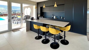 Eine moderne Küche mit einer zentralen Kochinsel, Barhocker, Pendelleuchten und Blick auf einen Pool durch große Schiebetüren.