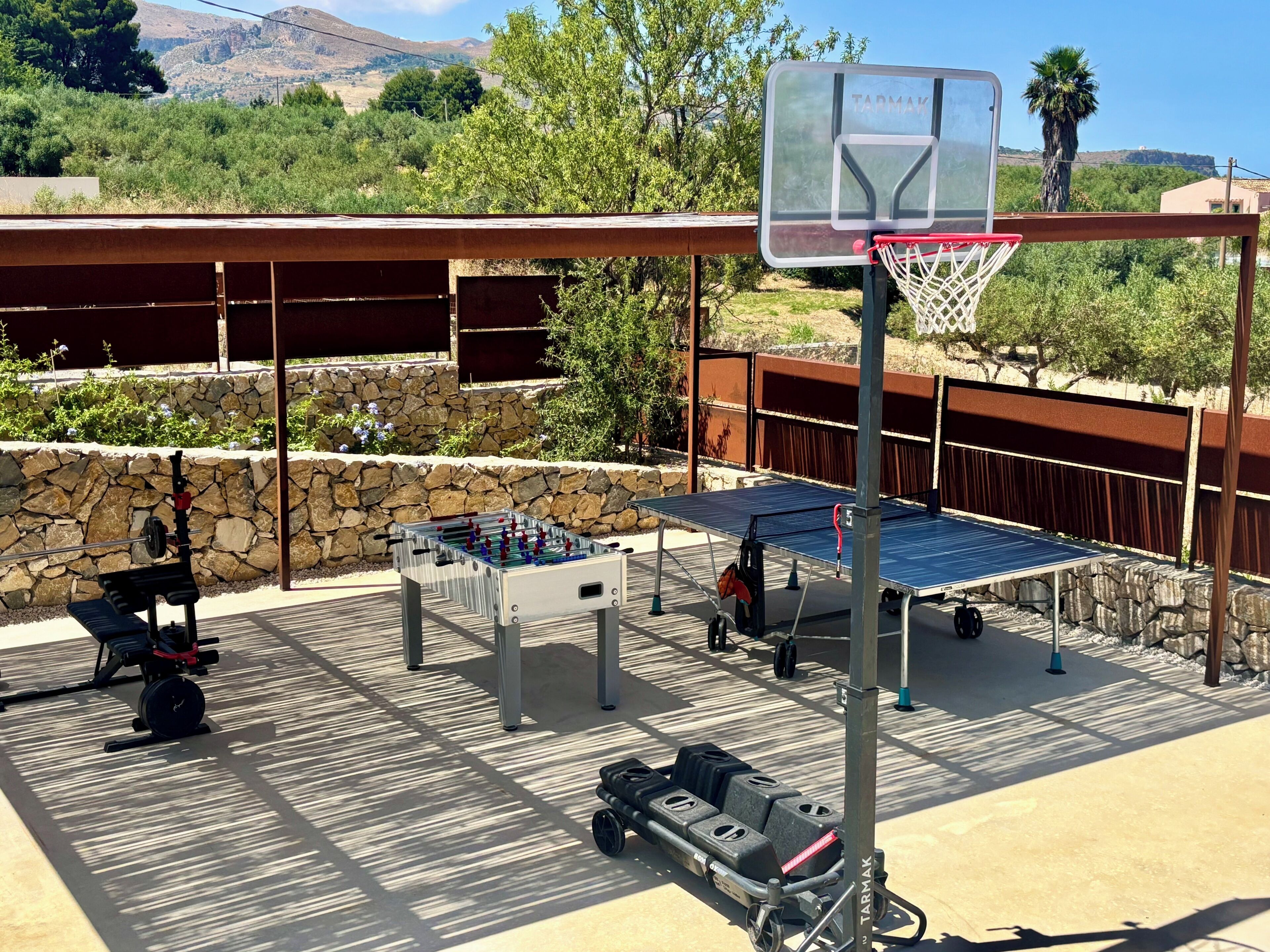An outdoor recreational area with a foosball table, a basketball hoop, and a table tennis table.