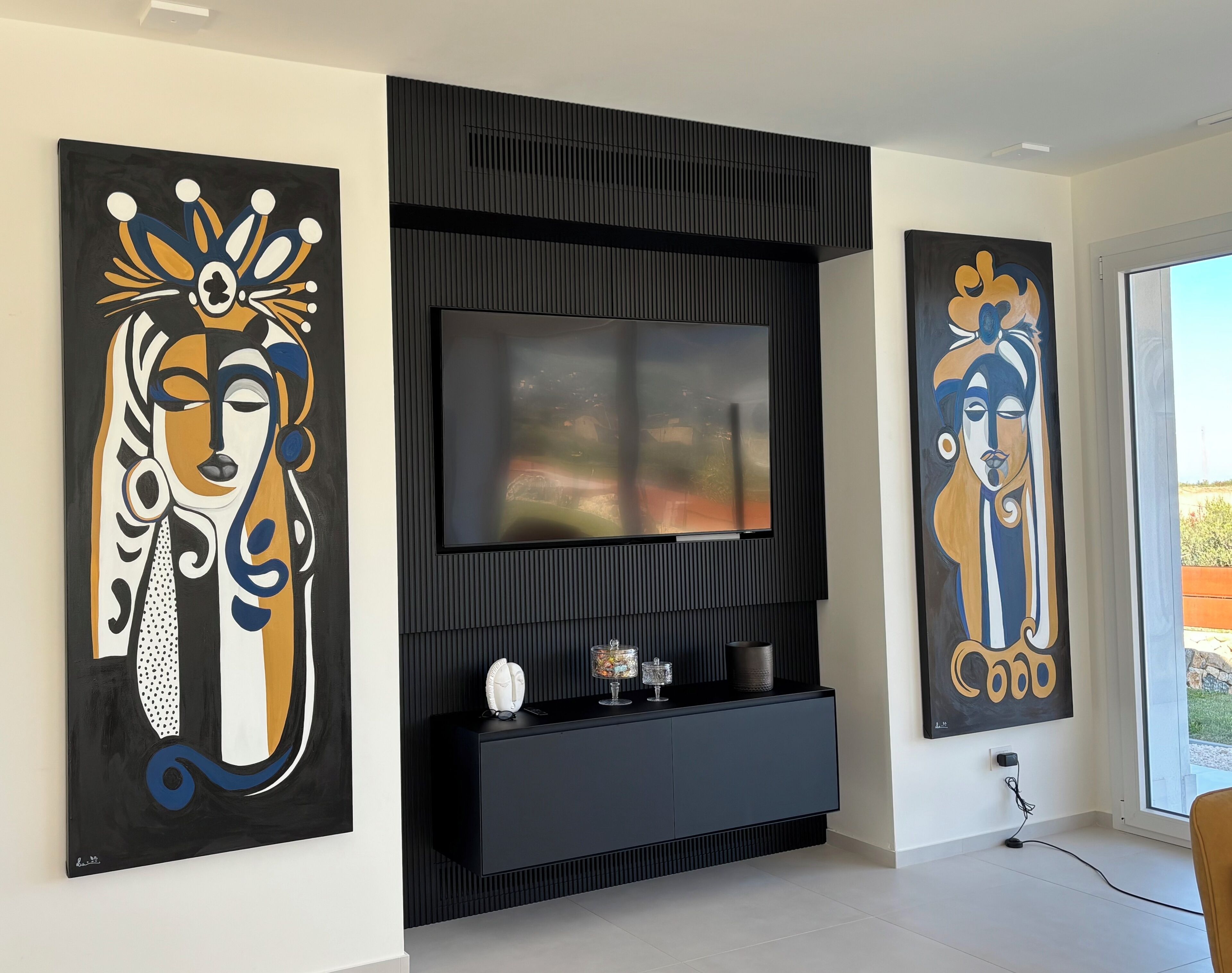 A modern living room with a large flat-screen TV mounted on a black wall, flanked by two large abstract paintings.