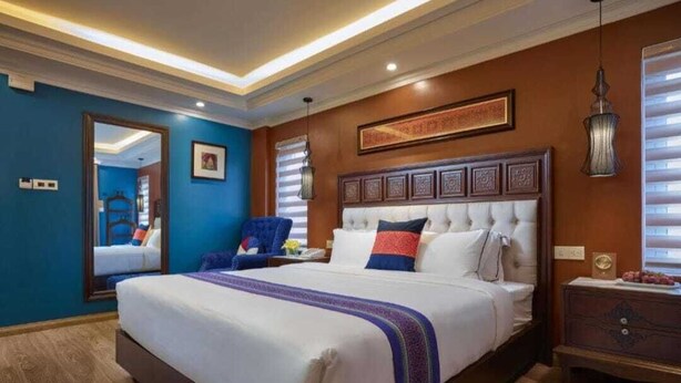 Een hotelkamer met een groot bed, een houten dressoir, een blauwe fauteuil en een kunstwerk in een lijst boven het bed.