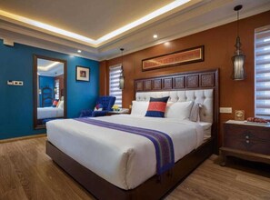 Een hotelkamer met een groot bed, een houten dressoir, een blauwe fauteuil en een kunstwerk in een lijst boven het bed.