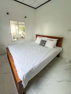 A bedroom with a wooden bed frame, white bedding, and a window with a view of outdoor plants.