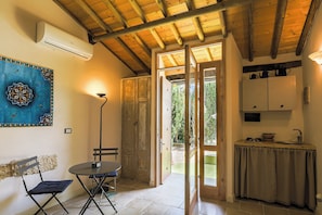 Ein Zimmer mit Holzboden, einem Esstisch mit Stühlen, einer Küchenzeile mit Schränken und Blick ins Freie durch eine offene Tür.